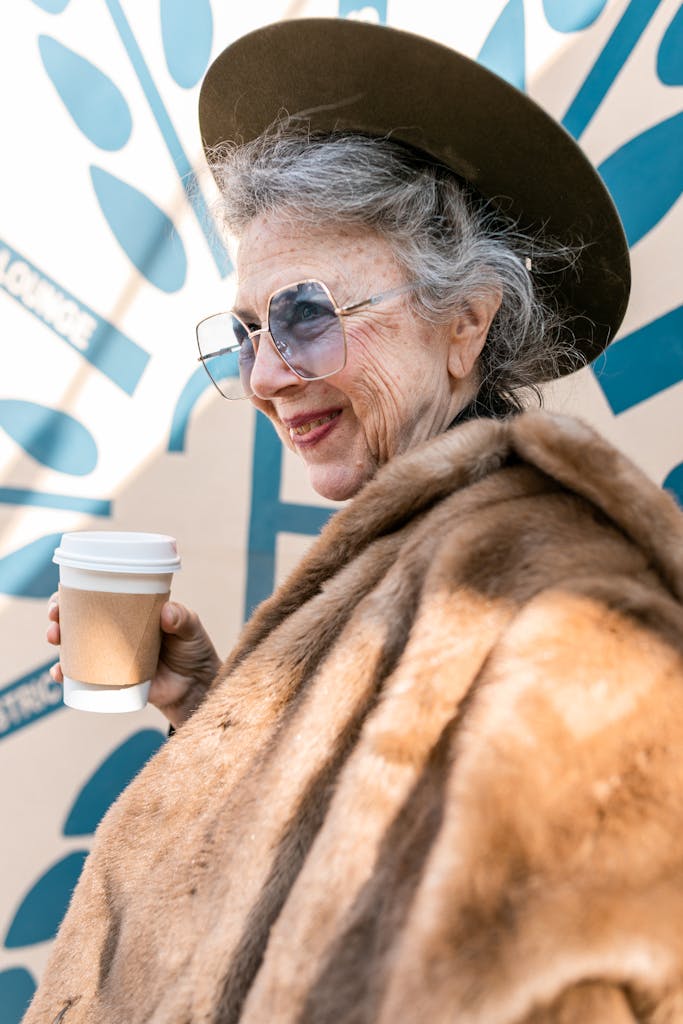 Elegant senior woman enjoying coffee outdoors, wearing a fur coat and hat, smiling.