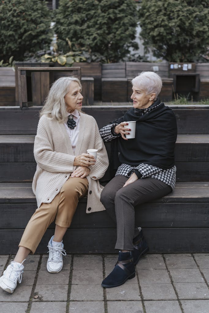 Two senior women sitting on a bench, sharing coffee and conversation in a cozy outdoor setting.