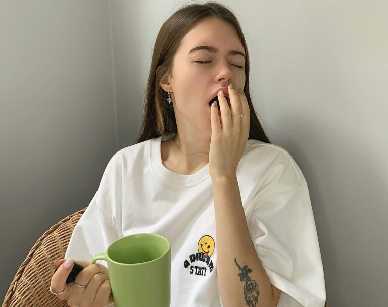 A young woman sits yawning in a chair, holding a green mug, dressed casually in a white shirt.