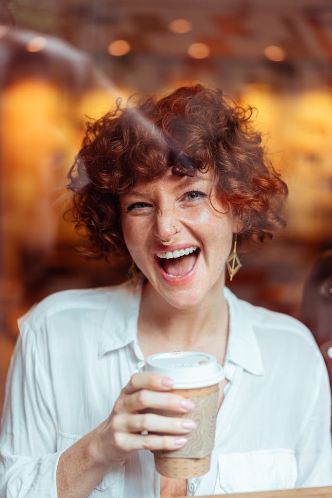 Smiling woman with curly red hair and coffee cup, captured in a cozy café environment.