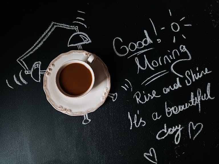 Cup of coffee on chalkboard with a cheerful good morning message and illustrations.