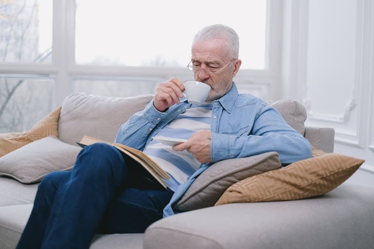 Elderly man enjoying a peaceful moment on sofa with a hot drink and book.