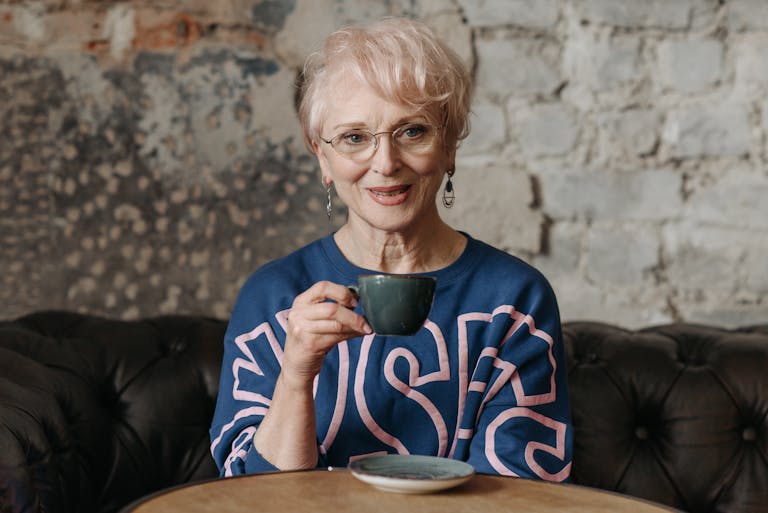 Senior woman with glasses enjoying a cup of coffee in a cozy cafe setting. Relaxed and content atmosphere.