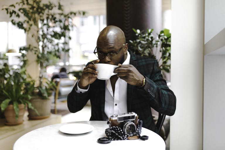 A man sipping coffee in a cafe with a vintage camera on the table. Relaxed indoor setting.