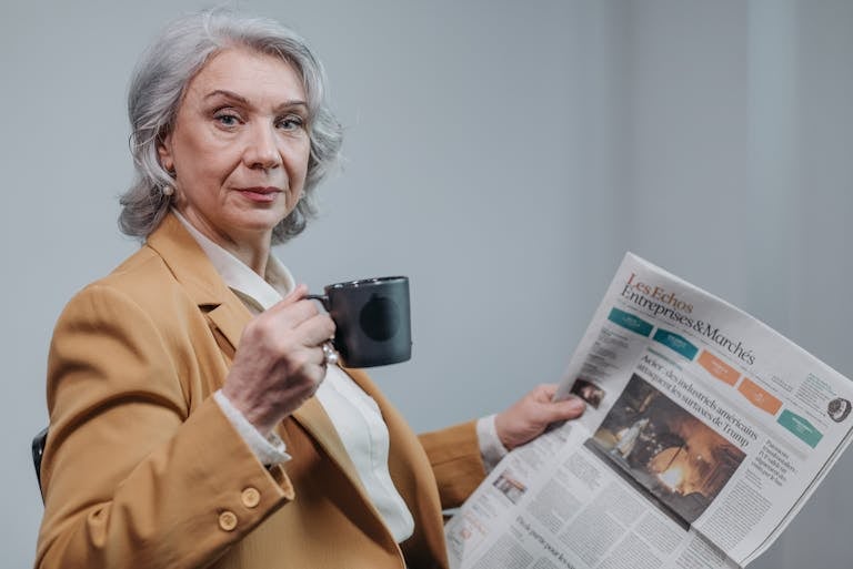 Confident senior businesswoman holding coffee and reading a newspaper indoors.