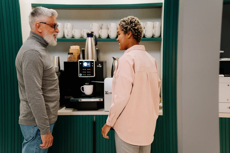 Senior and adult colleagues enjoying a coffee break, chatting by the coffee machine in an office break room.
