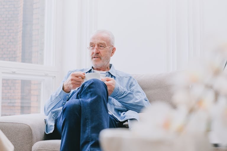 Senior man sipping tea on a cozy sofa in a bright, airy room with large windows.
