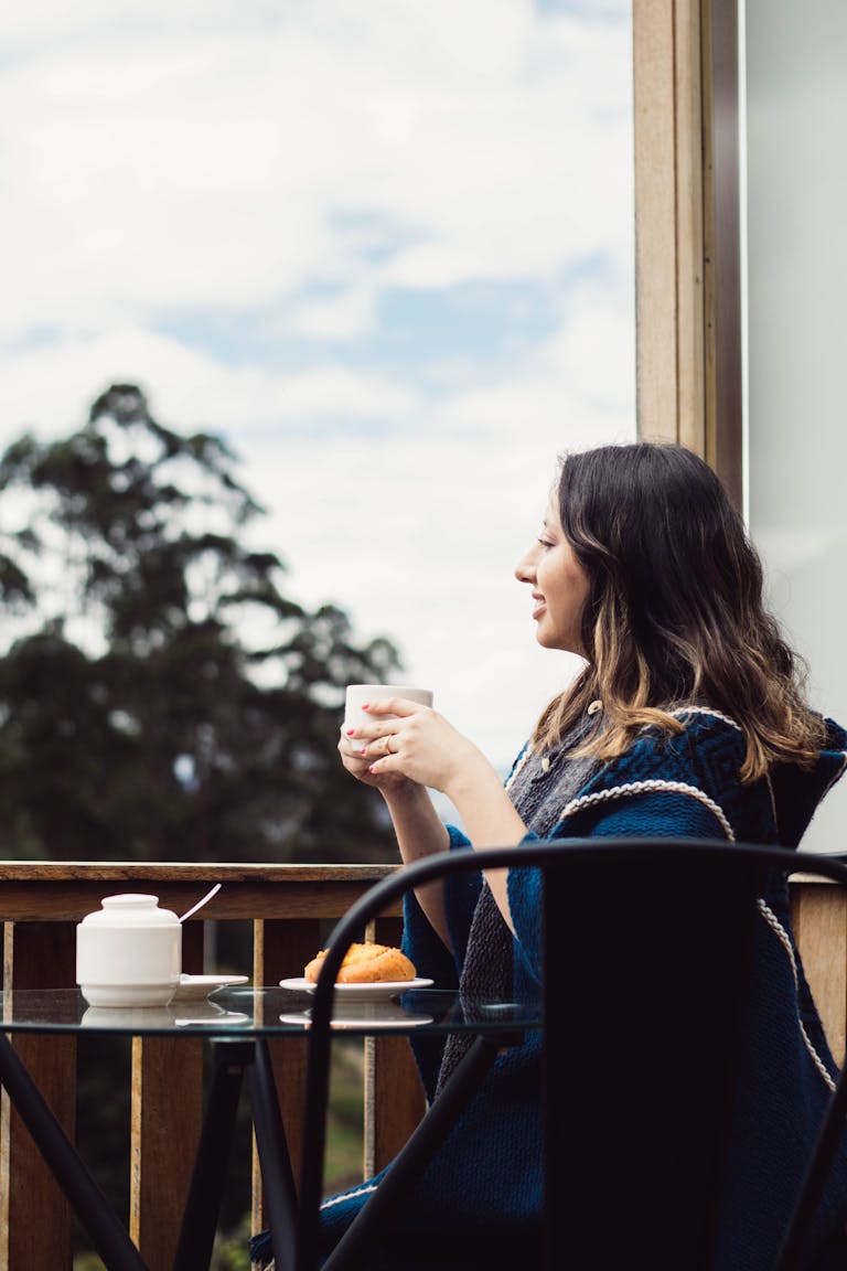 Woman wrapped in a blanket enjoying a warm drink on a balcony overlooking nature.