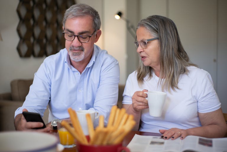 Senior couple relaxing with coffee, discussing smartphone content indoors. Warm and intimate moment.